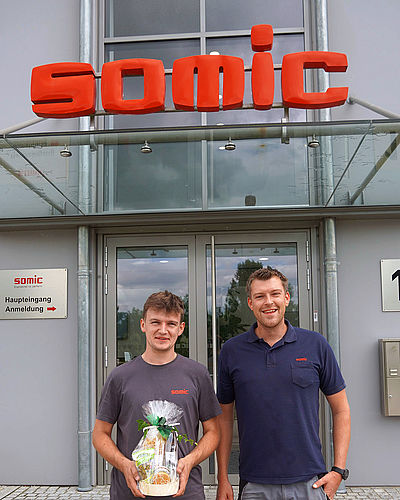 Ausbilder und Azubi mit Staatspreis vor SOMIC Haupteingang